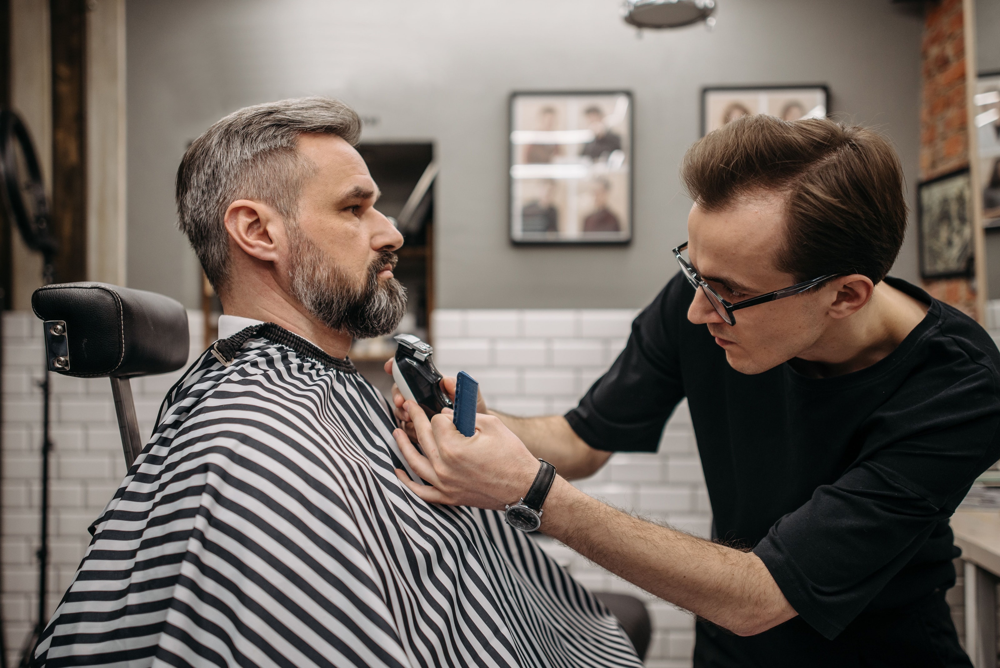 A barber trimming a customer's beard in a bright barbershop